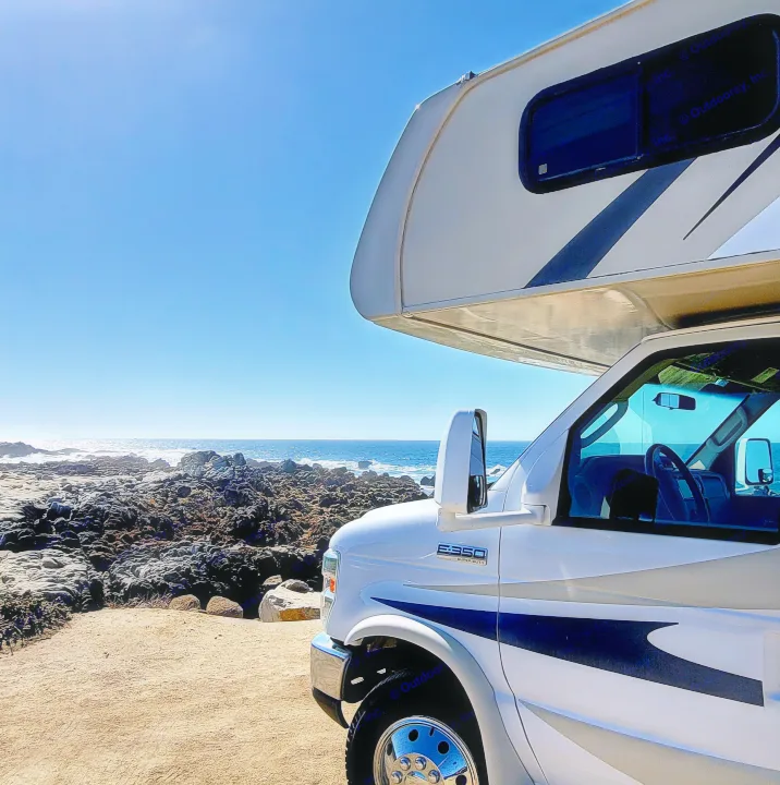 Experience coastal beauty! An RV rental is parked on a sandy patch beside rugged ocean rocks and blue waves under a clear sky.. Coachmen Leprechaun 2016