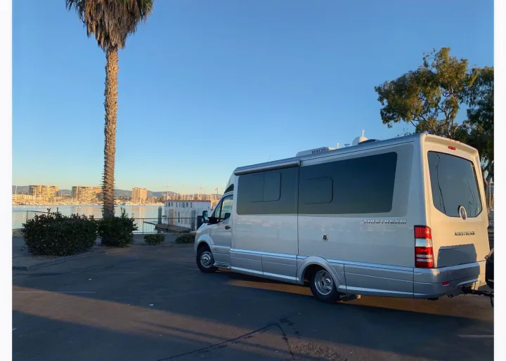 Silver Airstream RV rental van by a sunny marina with boats and buildings. A palm tree stands tall under a clear blue sky.. Airstream Atlas 2021