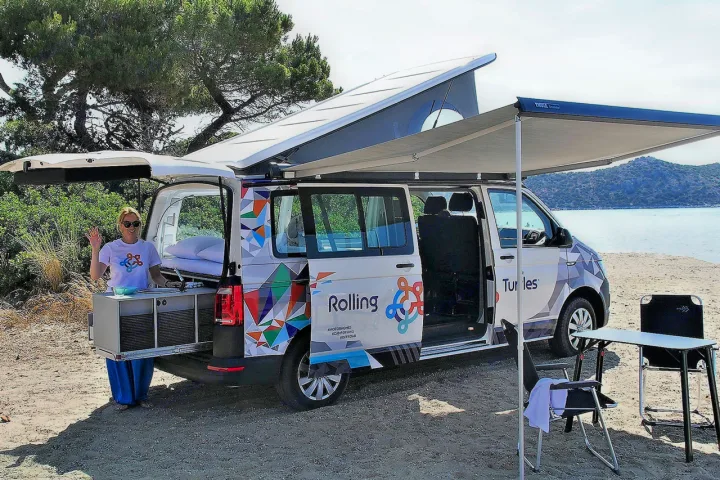 RV rental campervan with pop-top roof, awning, and pull-out kitchen on a beach. A woman waves.. Volkswagen T6 2020