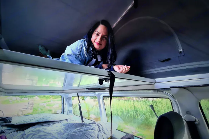 Woman smiles from a pop-top bunk in an RV rental, overlooking a cozy lower bed and green outdoor views.. Volkswagen T6 2020