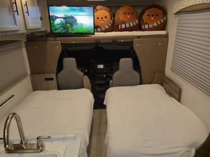 Inside an RV rental, showing two beds, a sink, and an overhead bunk with a TV and Chewbacca plushies near the cab.. Coachmen Freelander 2021