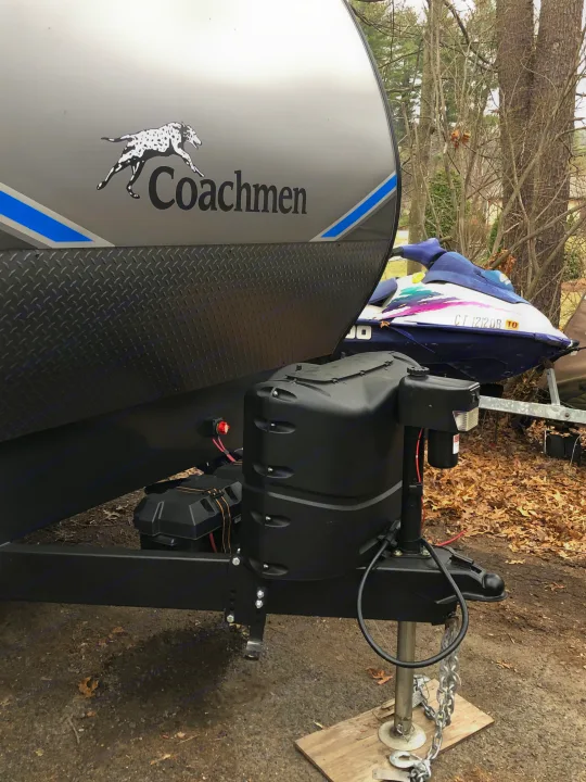 Coachmen RV rental travel trailer, showing front propane tanks, battery, and hitch. A jet ski is in the background.. Coachmen Catalina 2021