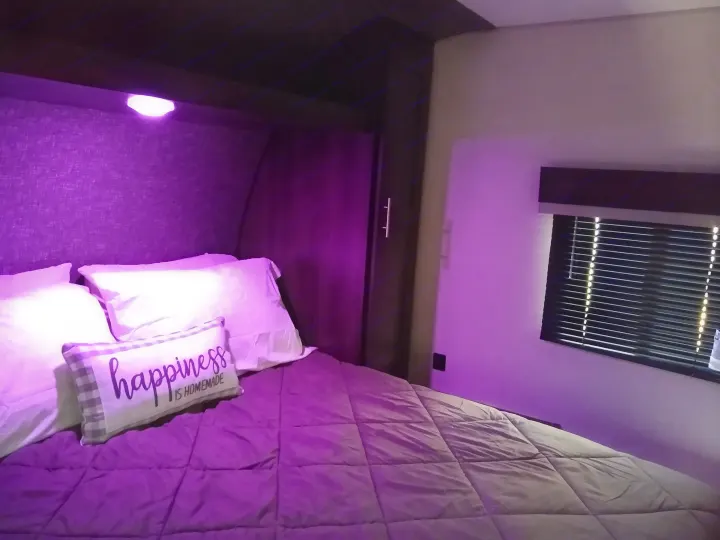 Cozy RV rental bedroom: bed with white pillows, purple comforter, and "happiness is homemade" pillow under purple ambient light.. KZ Sportsmen 2021