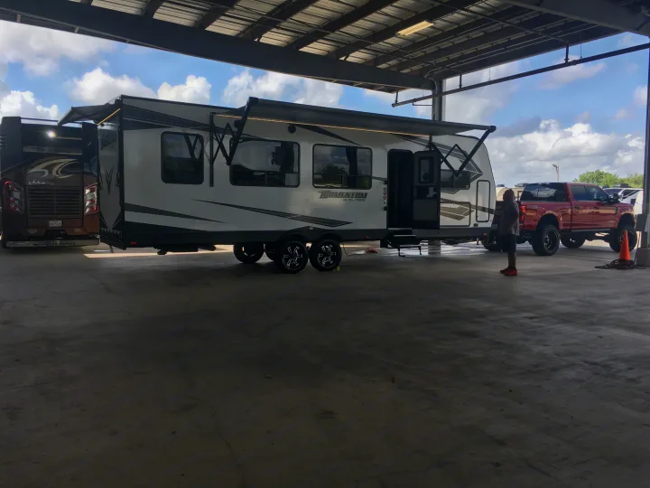 A white travel trailer with awning, a brown motorhome, and a red pickup truck are parked under a metal structure, possibly for RV rental service.. Grand Design Momentum 2020