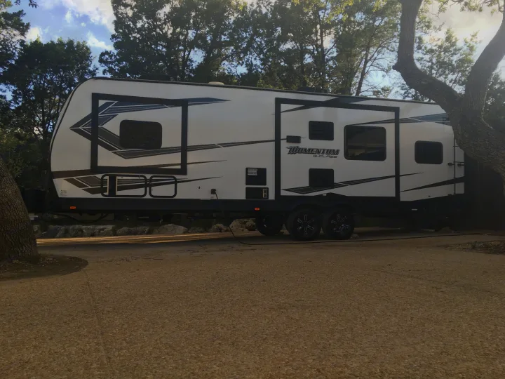 A white Grand Design Momentum G-Class travel trailer, an RV rental, parked and powered up among trees.. Grand Design Momentum 2020