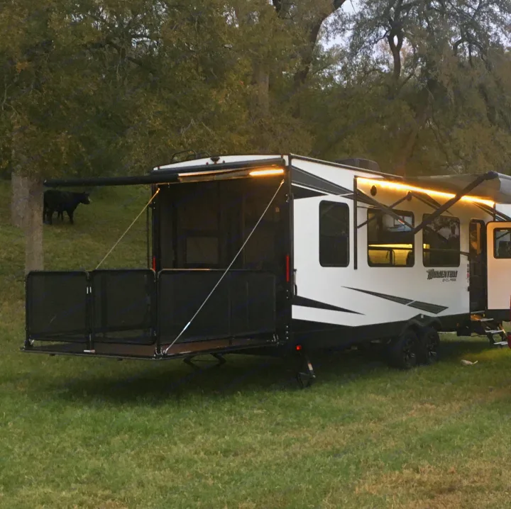 White RV rental set up with an extended deck and bright lights in a grassy field, with trees and a cow visible in the background.. Grand Design Momentum 2020