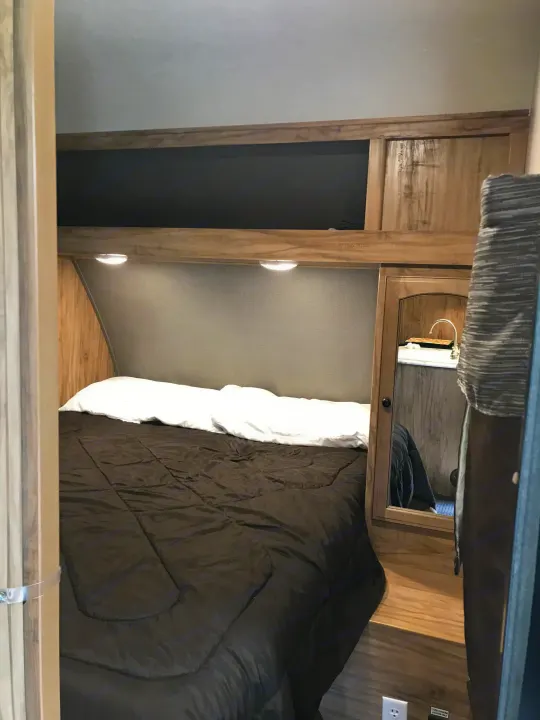 Cozy RV rental bedroom featuring a dark bed, overhead lights, wood cabinetry, and a mirrored sink area.. Coachmen Freedom Express 2018