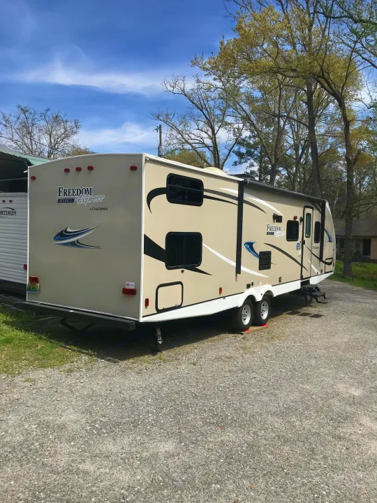 Tan Freedom Express travel trailer, an excellent RV rental option, parked on gravel under a blue sky, surrounded by trees.. Coachmen Freedom Express 2018