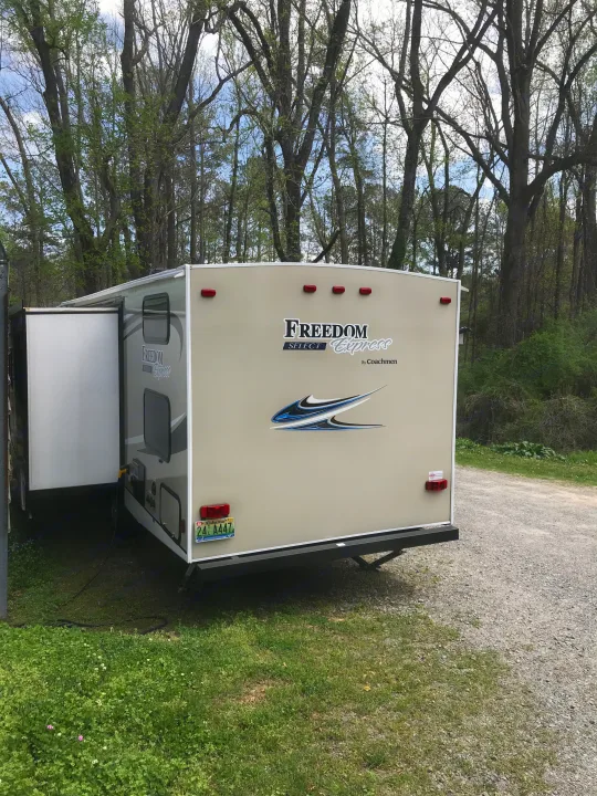 A beige Freedom Express RV rental travel trailer's rear view, with an Alabama plate, parked on gravel amidst spring trees.. Coachmen Freedom Express 2018