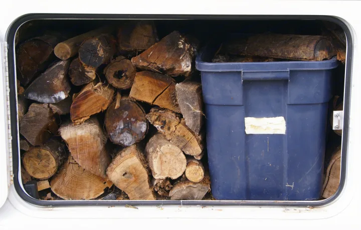 An RV rental storage compartment is packed with various sized firewood logs and a large blue plastic bin.. Winnebago Vista 2013