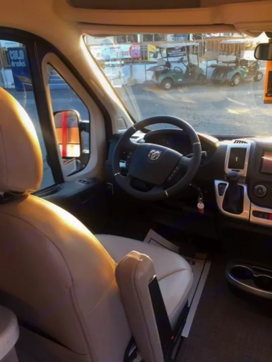 Driver's perspective inside an RV rental: Ram ProMaster cab with dashboard, seat, and steering wheel. Golf carts visible outside.. Thor Sequence 2021