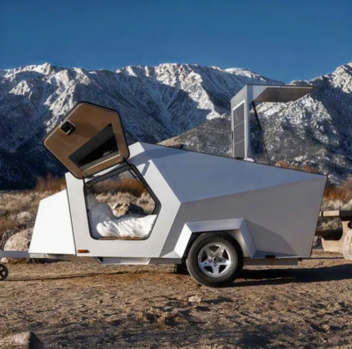 A futuristic, angular travel trailer, perfect for an RV rental, is parked among snow-capped mountains. A bed is visible inside.. Polydrop K-20 2020