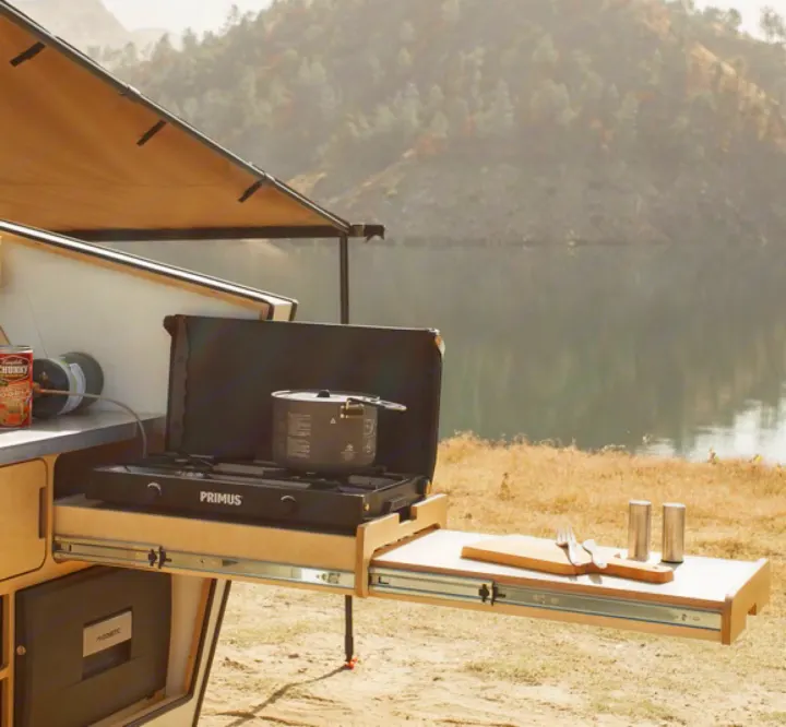 An RV rental features an outdoor kitchen with a camping stove, pot, and prep area by a scenic lake, perfect for outdoor cooking.. Polydrop K-20 2020
