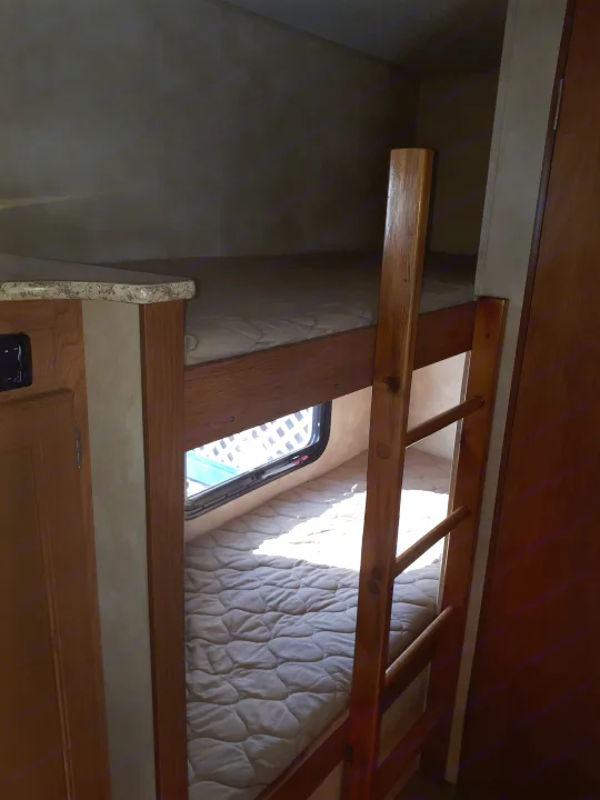 Inside an RV rental, showing a set of wooden bunk beds with quilted mattresses and a window letting in sunlight.. Coachmen Clipper 2013