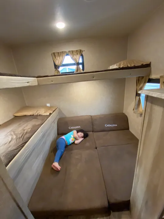 Cozy RV rental interior shows child asleep on a Catalina sofa bed below bunk beds. Ideal for family travel adventures.. Coachmen Catalina 2019
