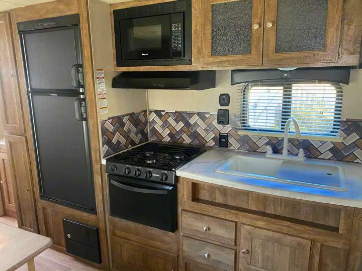RV rental kitchen with black appliances: fridge, microwave, Suburban stove, and sink. Wood cabinets & herringbone backsplash.. Forest River Puma 2019