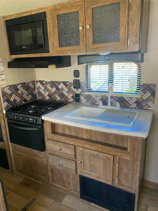 RV rental kitchen featuring a microwave, stove, sink, cabinets, and herringbone backsplash.. Forest River Puma 2019