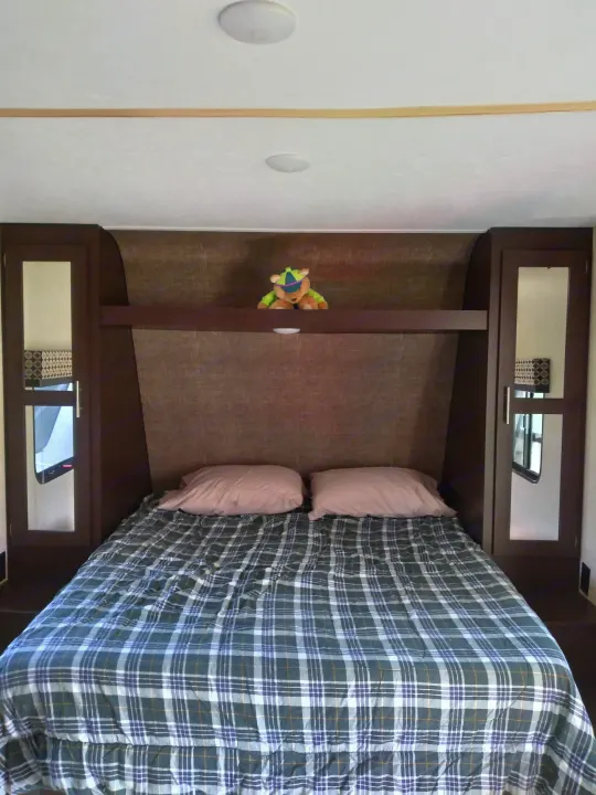 Cozy RV rental bedroom with a plaid bed, pink pillows, and built-in dark wood cabinets. A stuffed animal rests on the shelf.. KZ Other 2017