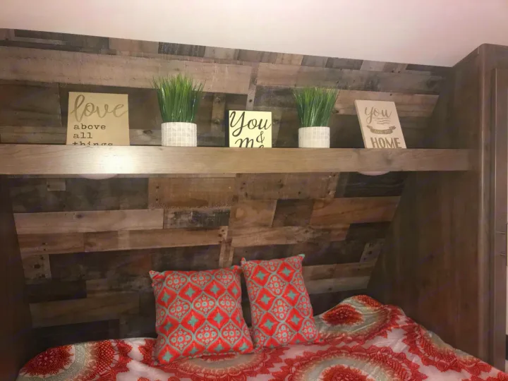 Inside an RV rental: a cozy bedroom with a planked wood headboard, decorative shelf, plants, signs & colorful bedding.. Dutchmen Aspen Trail 2021