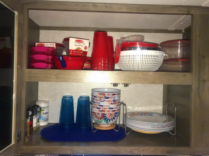 Organized kitchen cabinet in an RV rental, featuring plastic storage, colorful dinnerware, and cups, optimizing space.. Dutchmen Aspen Trail 2021