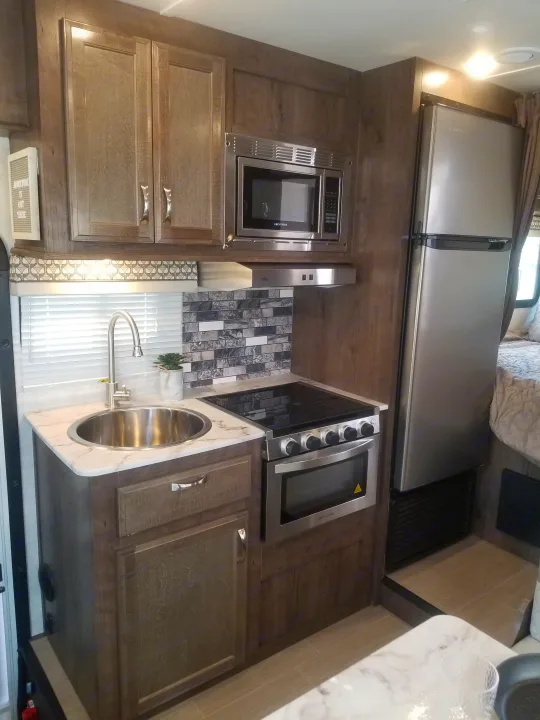 Modern compact kitchen in an rv rental, with stainless steel appliances, marble-look counters, and wood cabinetry.. Gulf Stream Conquest 2021