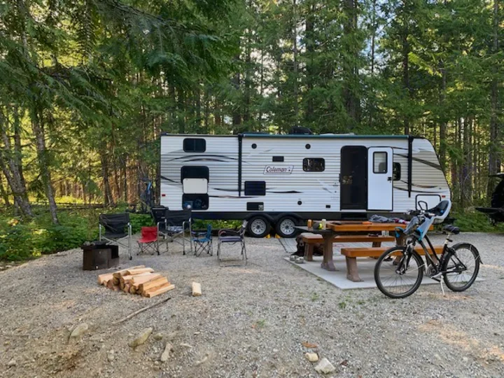 An rv rental trailer parked in a gravel forest campsite with a picnic table, fire pit, chairs, and bicycle.. Coleman Lantern Edition 262 BH 2017