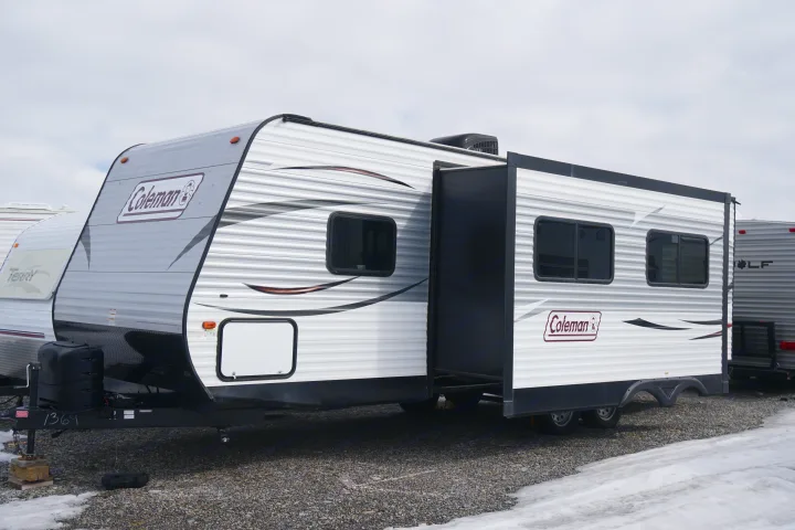 A white Coleman travel trailer RV rental, featuring an extended slide-out, is parked on a gravel lot with patches of snow.. Coleman Lantern Edition 262 BH 2017