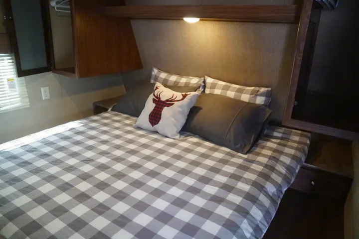RV rental bedroom featuring a cozy bed with gray buffalo plaid bedding, a deer accent pillow, and overhead storage.. Coleman Lantern Edition 262 BH 2017