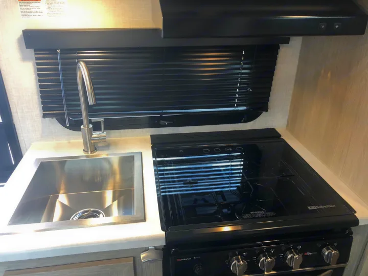 RV rental kitchen: Stainless steel sink, gooseneck faucet, black Suburban stove/oven, range hood, and window with blinds.. Forest River Rockwood Geo Pro 2021