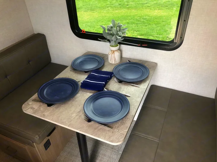 Cozy RV rental dinette with set table for four, faux plant, and a view of green fields through the window.. Forest River Rockwood Geo Pro 2021