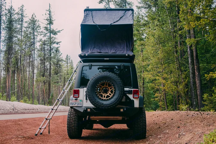 Outdoor rv rental: A white Jeep Wrangler, equipped with a rooftop tent and ladder, parked on a forest trail for camping.. Jeep Wrangler Rubicon 2017