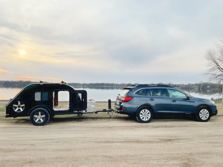 RV rental: Subaru Outback towing a black teardrop camper by a lake. Perfect for serene outdoor travel.. American Teardrop Travel Trailer 2016