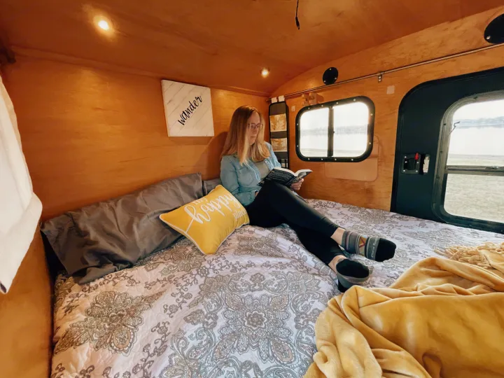 Enjoy a peaceful moment in your next RV rental. A woman reads on the comfortable bed inside a rustic camper with a scenic view outside the window.. American Teardrop Travel Trailer 2016