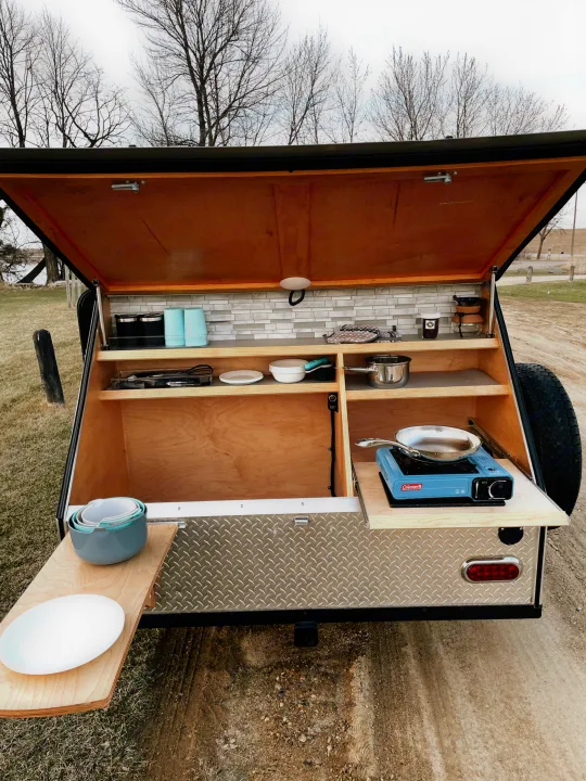 Compact outdoor kitchen in an RV rental teardrop trailer, set up for camping with stove, dishes, and pull-out counter.. American Teardrop Travel Trailer 2016