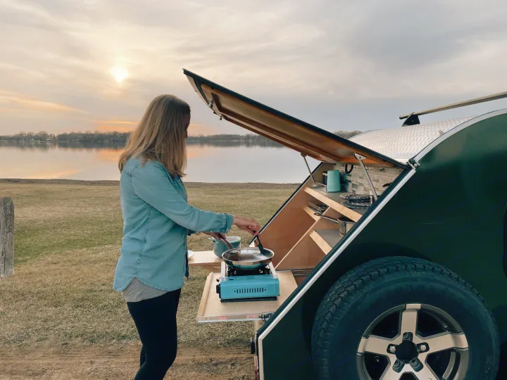 RV rental adventure: Woman cooking lakeside from her teardrop trailer's compact outdoor kitchen at sunset.. American Teardrop Travel Trailer 2016