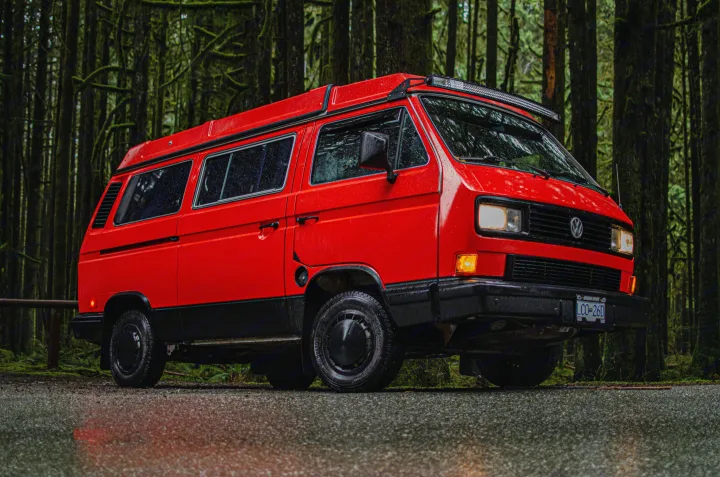 Bright red RV rental van parked on a wet road in a dark, mossy forest, ready for outdoor adventures.. Volkswagen Vanagon 1989