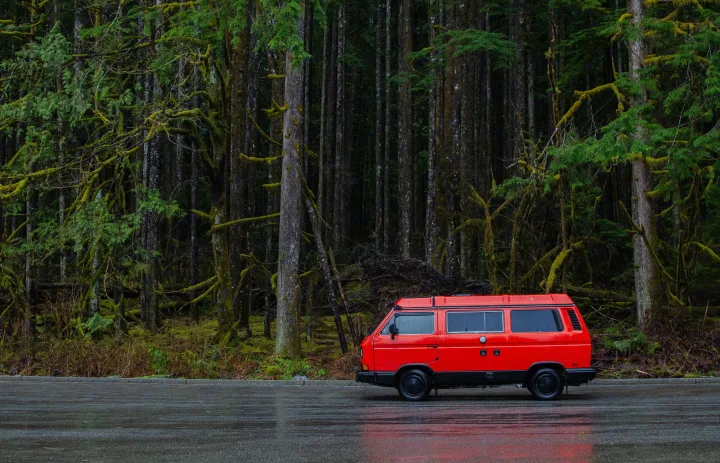 Red campervan RV rental parked on a wet road next to a dense, moss-covered forest. Ideal for a nature adventure.. Volkswagen Vanagon 1989