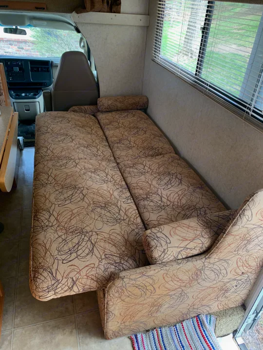 Inside an RV rental, a patterned convertible sofa bed stretches towards the driver's cab, with a window and blinds visible.. Winnebago Access 2007