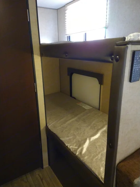 Inside an RV rental: a top bunk with a remote, lower bunk area, and bright window with blinds, showing compact sleeping space.. Forest River Cherokee Wolf Pup 2017
