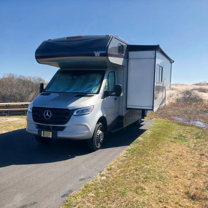 Silver Mercedes-Benz Sprinter RV rental with extended slide-out at a sunny campsite beside sandy dunes.. Winnebago Mercedes Sprinter 2020