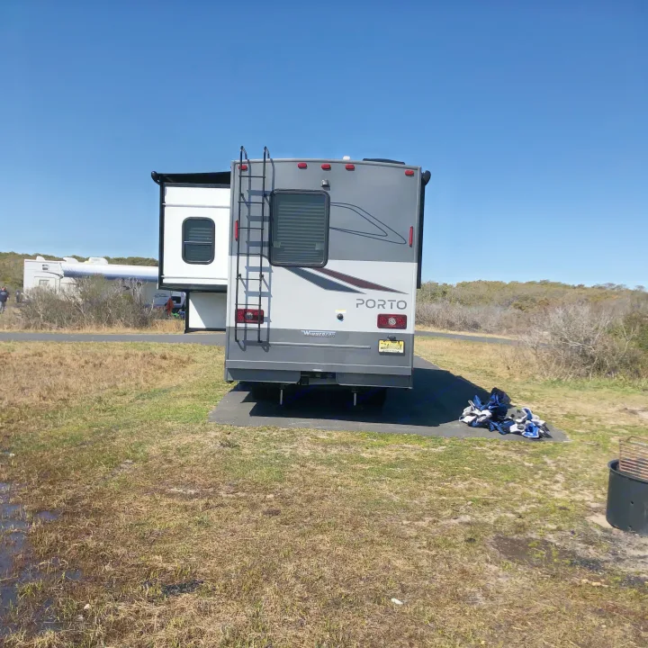 A grey Winnebago Porto RV rental is parked on an asphalt pad at a sunny campground site, with dry grass.. Winnebago Mercedes Sprinter 2020
