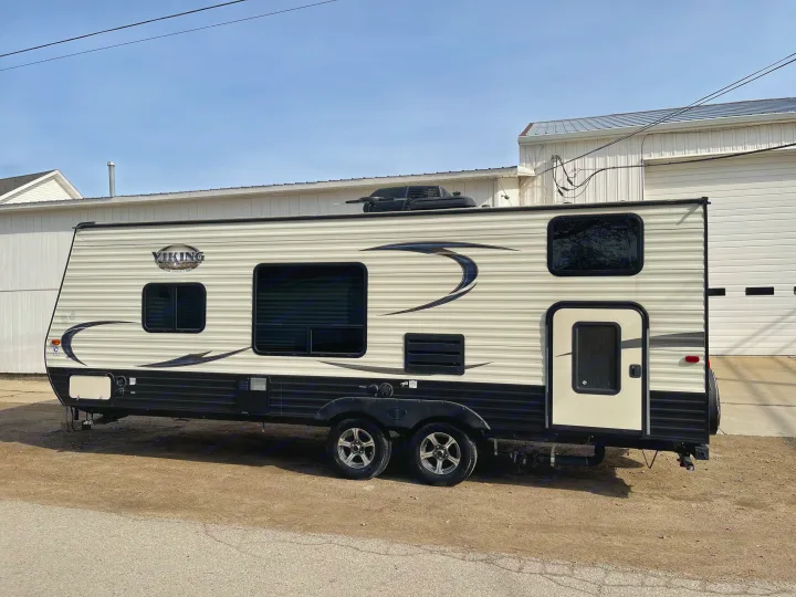 Beige Viking travel trailer, an RV rental option, parked on gravel with its door open under a clear sky.. Viking 21BH 2018