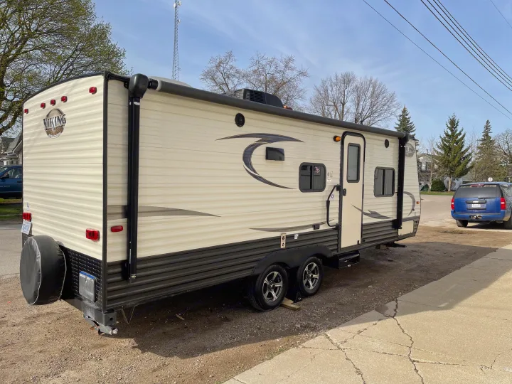 Light-colored Viking RV rental, parked on gravel with a sidewalk nearby. Shows side with awning, windows, rear spare tire, and dual axles.. Viking 21BH 2018