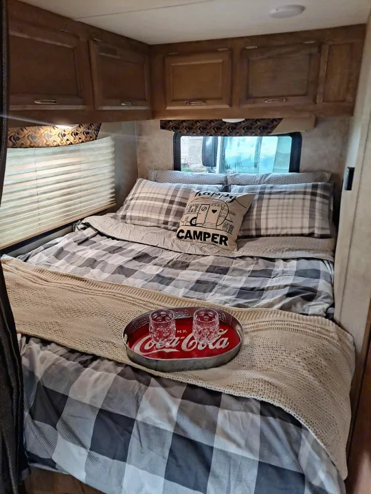 Inside a cozy RV rental bedroom: plaid bedding, a "happy CAMPER" pillow, and a Coca-Cola tray.. Gulf Stream Conquest 2018