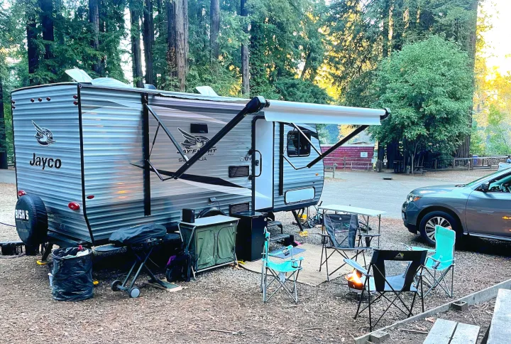 RV rental at a serene forest campsite. Jayco travel trailer with awning, camping chairs, and grill ready for a relaxing outdoor evening.. Jayco Jay Flight 2021