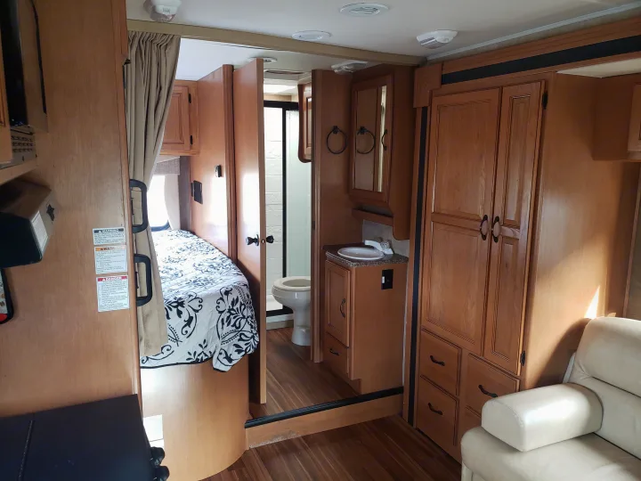 This RV rental interior showcases a cozy bedroom with a patterned bedspread, a compact bathroom with a toilet and sink, and functional storage cabinets.. Coachmen Prism 2150LE 2013