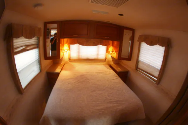 A cozy RV rental bedroom interior with a comfortable bed, wood cabinetry, and bedside lamps, perfect for your journey.. Winnebago Adventurer 1999