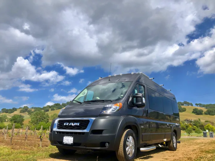 Dark gray Ram ProMaster camper van rv rental, parked in a vineyard, under a partly cloudy sky.. Jayco Swift 2021