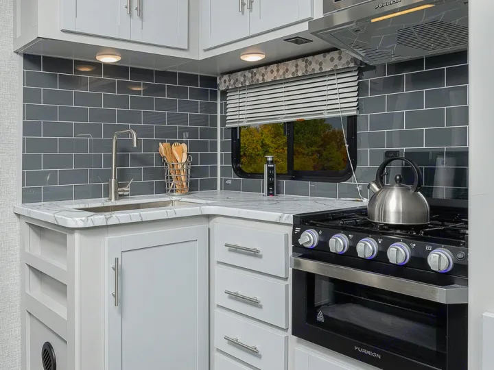 This modern RV rental kitchen has gray subway tile, marble counters, a Furrion range, and a window overlooking autumn trees.. Other Bullet 2021