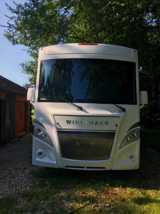 Front view of a sleek, white Winnebago RV rental with chrome grille and headlights, parked on gravel near trees.. Winnebago Intent 2019